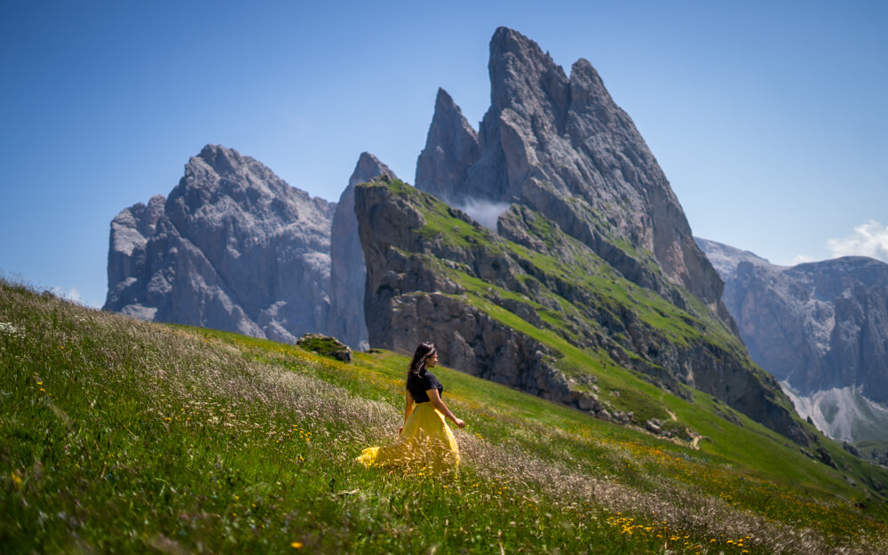 Girl standing in the meadows full of wildflowers commonly seen in the Dolomites in July. In the backgroud are the jagged peaks of Seceda