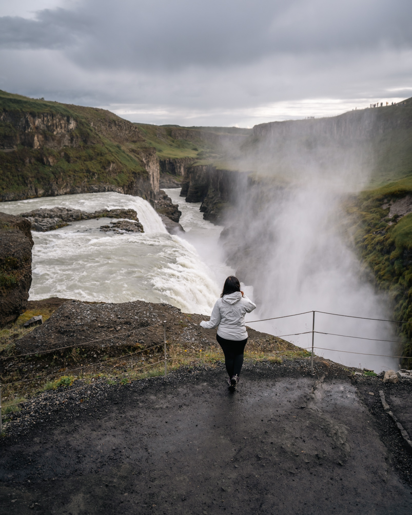 A girl standing infront of a waterfall in Iceland wearing a rain jacket which is essential on an Iceland summer packing list