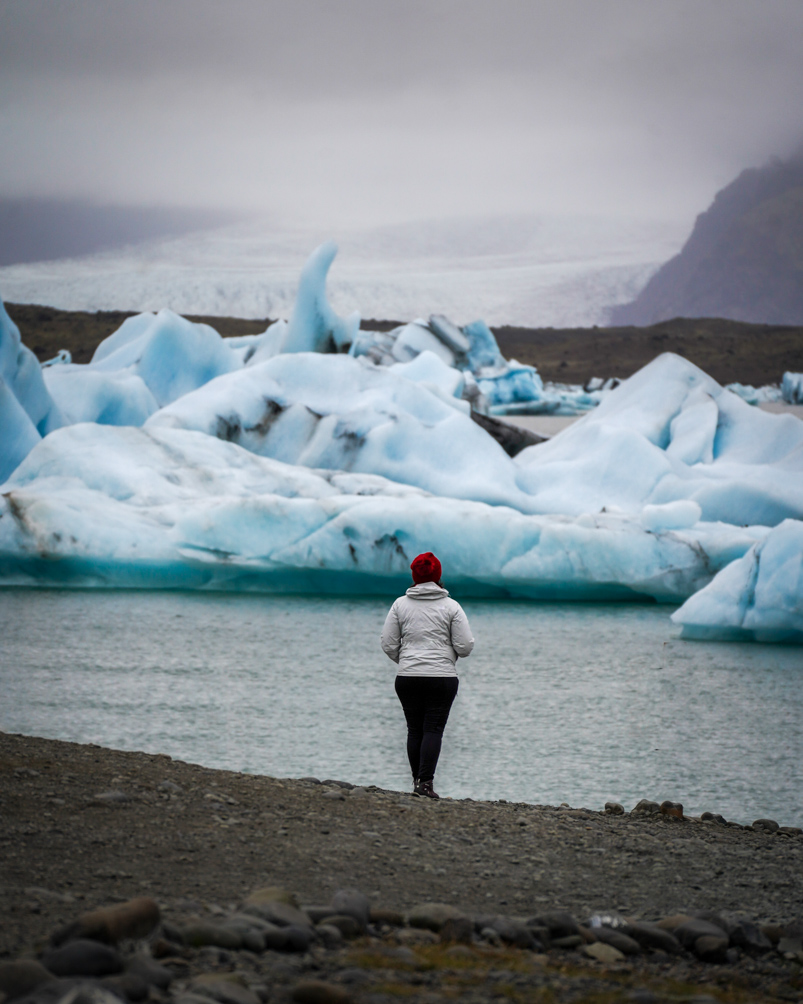 A girl in a white jacket and red beanie standing in front of a lagoon with giant icebergs. In the distance, you can even see a glacier.