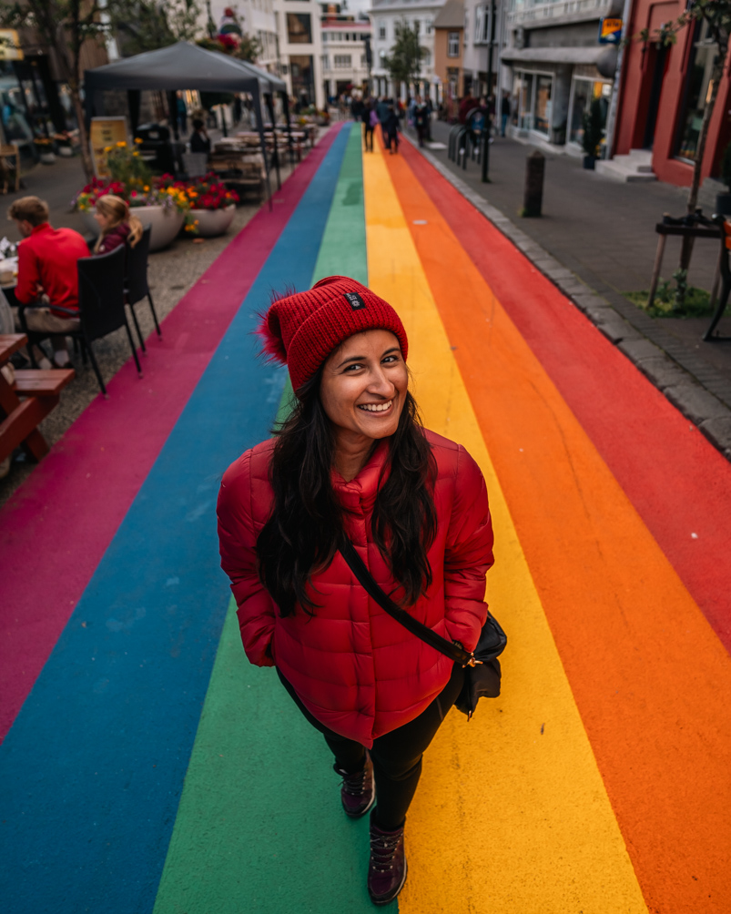 A girl posing for the camera wearing a maroon jacket and beanie standing over a rainbow coloured street