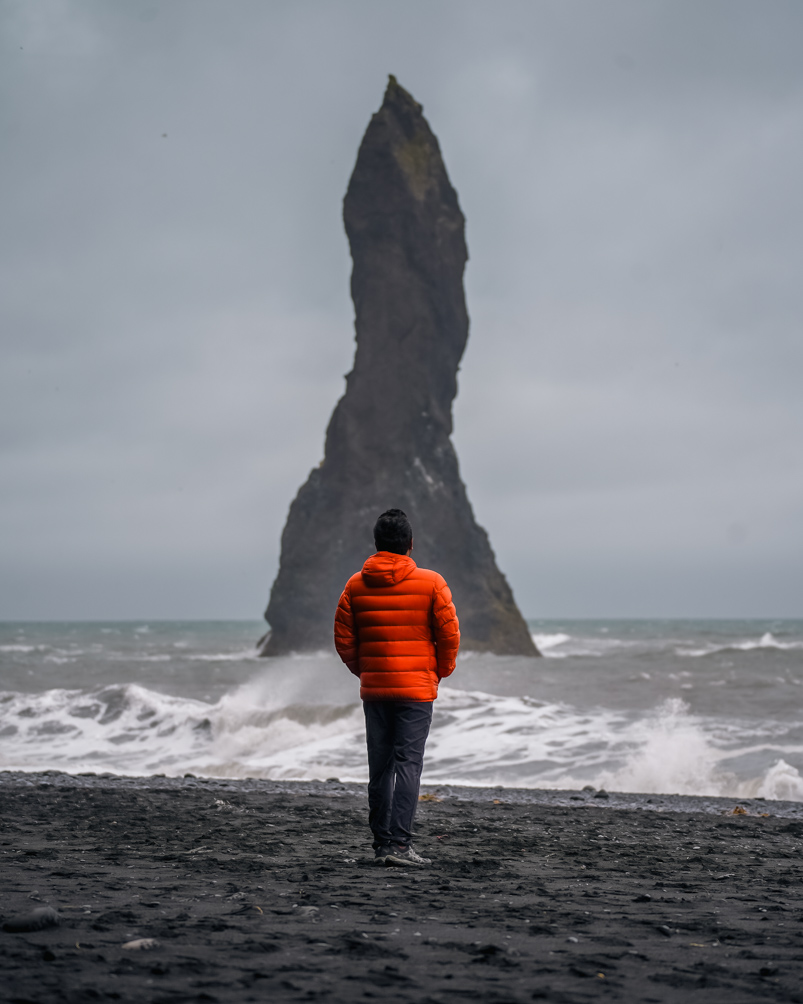 A man standing on a black sand beach looking at the sea with a giant rock protruding from it.