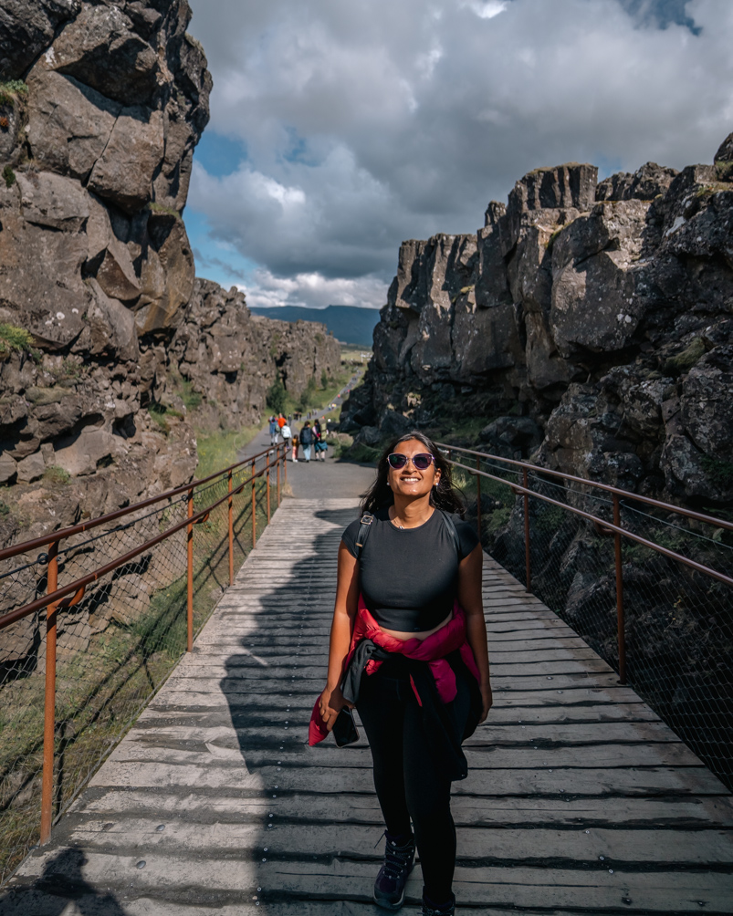 A girl walking between tectonic plates in Thingvellir National Park in Iceland wearing a black t-shirt and pants with her sweaters and jackets tied at the waist. Layers are essential for an Iceland summer packing list!