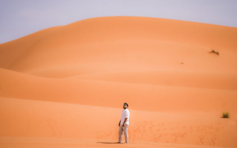 A man stands in the desert looking up with a background of layers of towering sand dunes