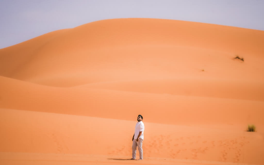 A man stands in the desert looking up with a background of layers of towering sand dunes
