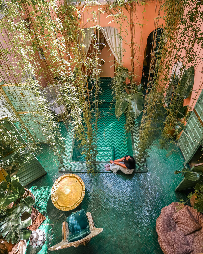 A girls sits by a pool of water surrounded by traditional Moroccan tiles and hanging plants in a riad in Marrakesh