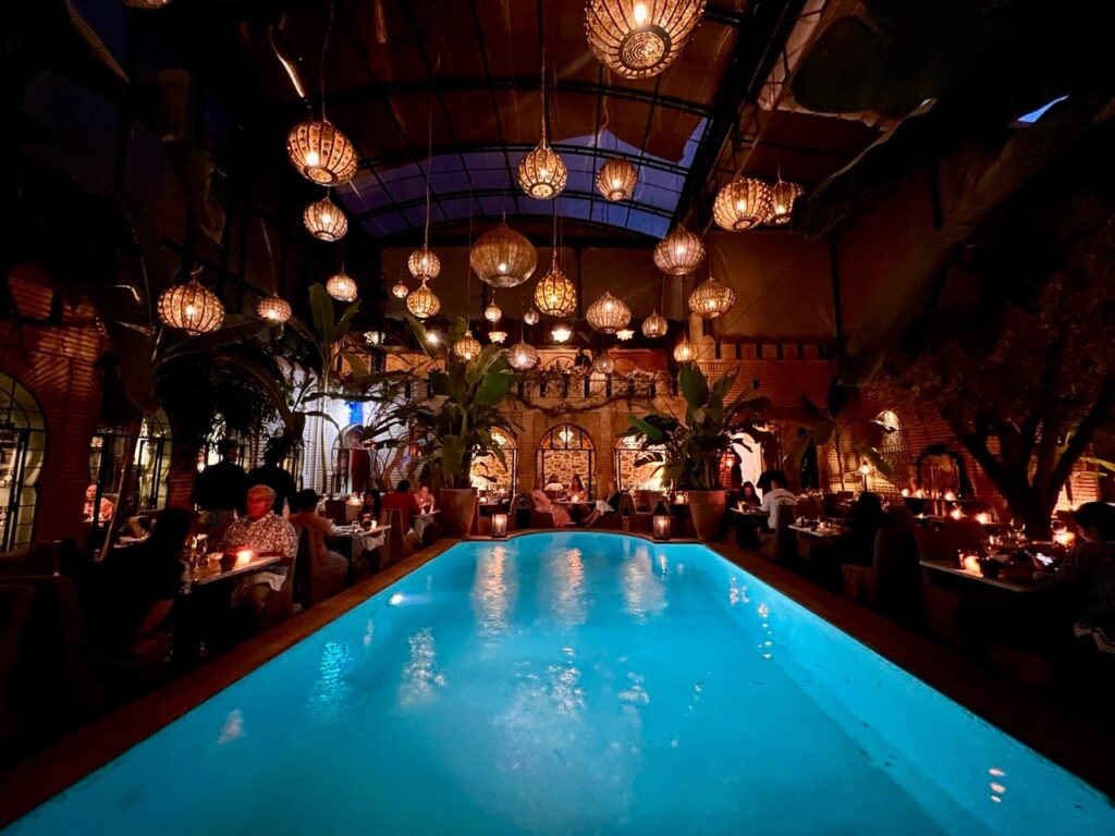 People sitting and eating by a pool with lanterns hanging above in a cosy ambience at La Trattoria restaurant in Marrakesh