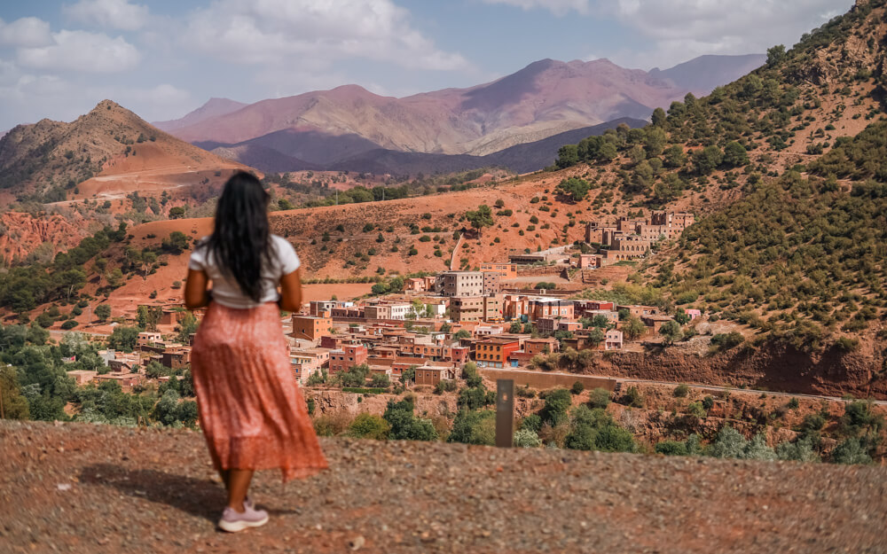 A woman in a flowing skirt gazes at a colorful village nestled among the reddish Atlas mountains and green trees under a partly cloudy sky.on her drive to Marrakesh in Morocco