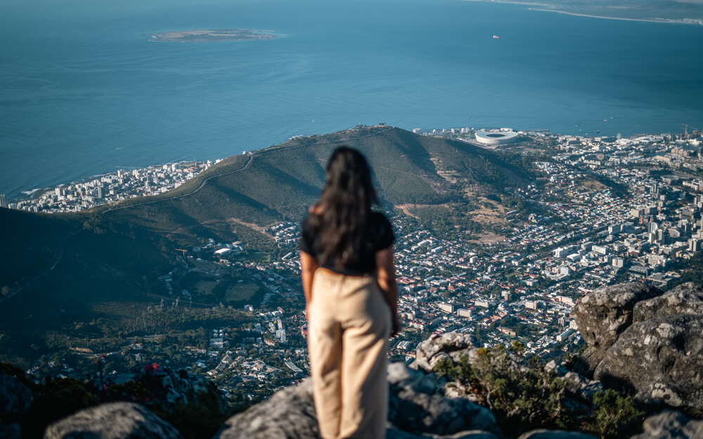 A woman stands on a rocky outcrop of Table Mountain (a must on this 3-day Cape Town Itinerary), overlooking Cape Town and the ocean, with a view of Signal hill the background.