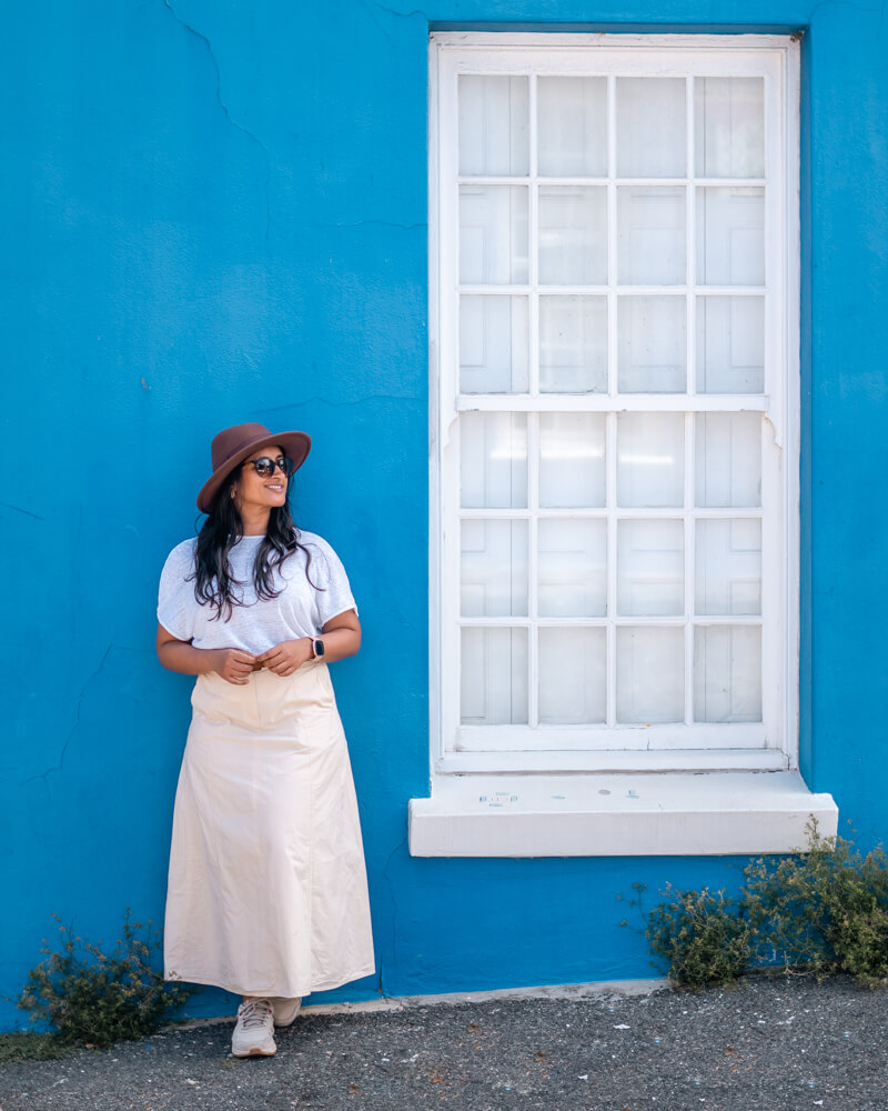 A woman stands against a vibrant blue wall in Bo-Kaap, wearing a white top and long cream skirt, next to a large white-framed window.