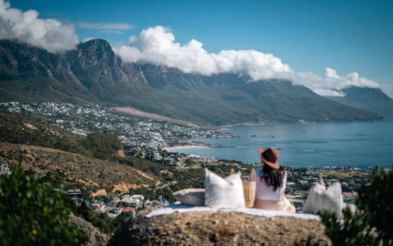 A girl sitting in a picnic style setup with a view of Cape Town's Twelve Apolstle from The Rock at Camps Bay