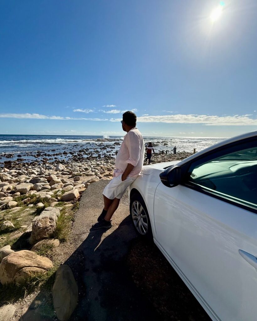 A man leans on a rented white car parked along the coast at Cape of Good Hope