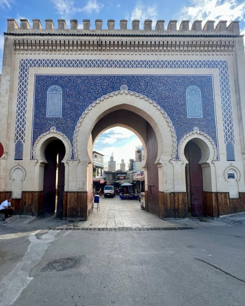 A grand archway adorned with intricate blue tiles leads into the  bustling markets of the Fes medina, framed by traditional Moroccan architecture.