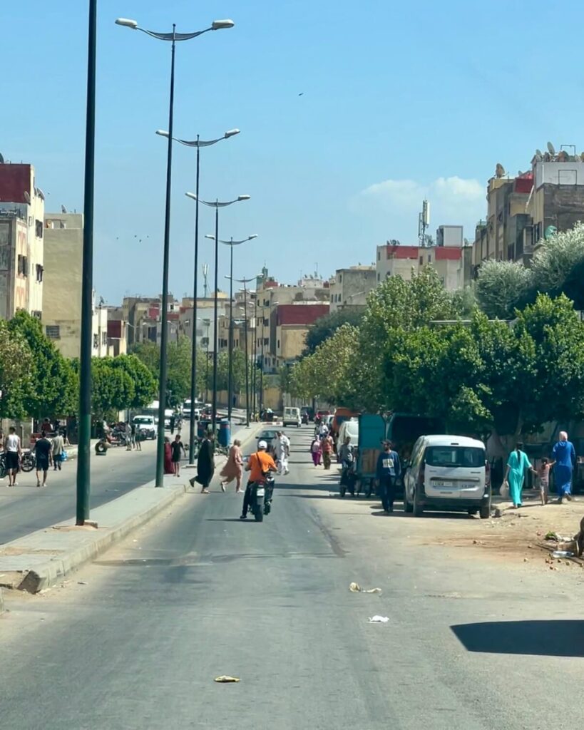 Busy street scene in Fes with pedestrians, motorcycles, and parked vehicles, flanked by trees and buildings under a clear blue sky.