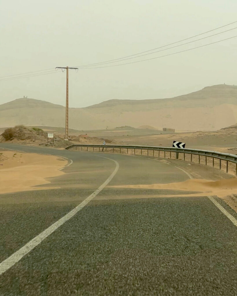 A winding road through a dusty desert landscape, with a power pole and sandy dunes in a hazy atmosphere!