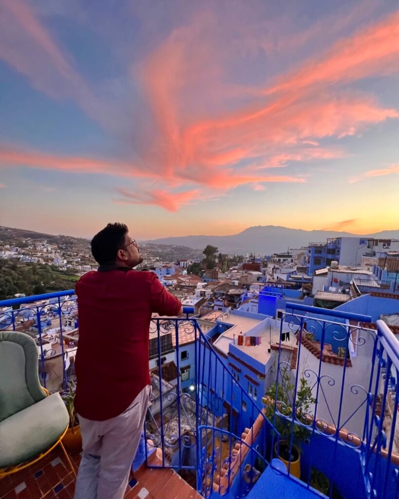 A person gazes out over the vibrant blue city of Chefchaouen at sunset, with colorful clouds illuminating the sky above. 