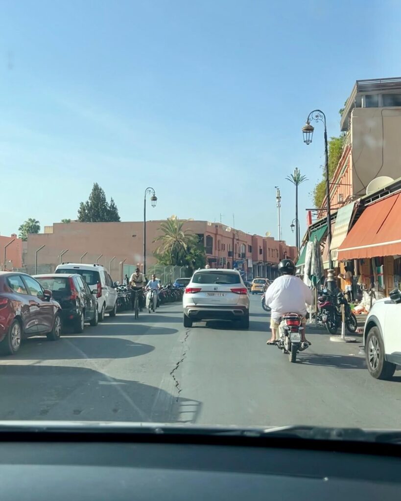 A busy street in Marrakech with cars, bicycles, and scooters, lined by shops under a clear blue sky. Palm trees dot the scene.