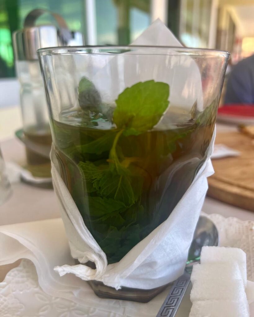 A glass of Moroccan mint tea with fresh mint leaves, wrapped in a napkin, sits on a table alongside sugar cubes.