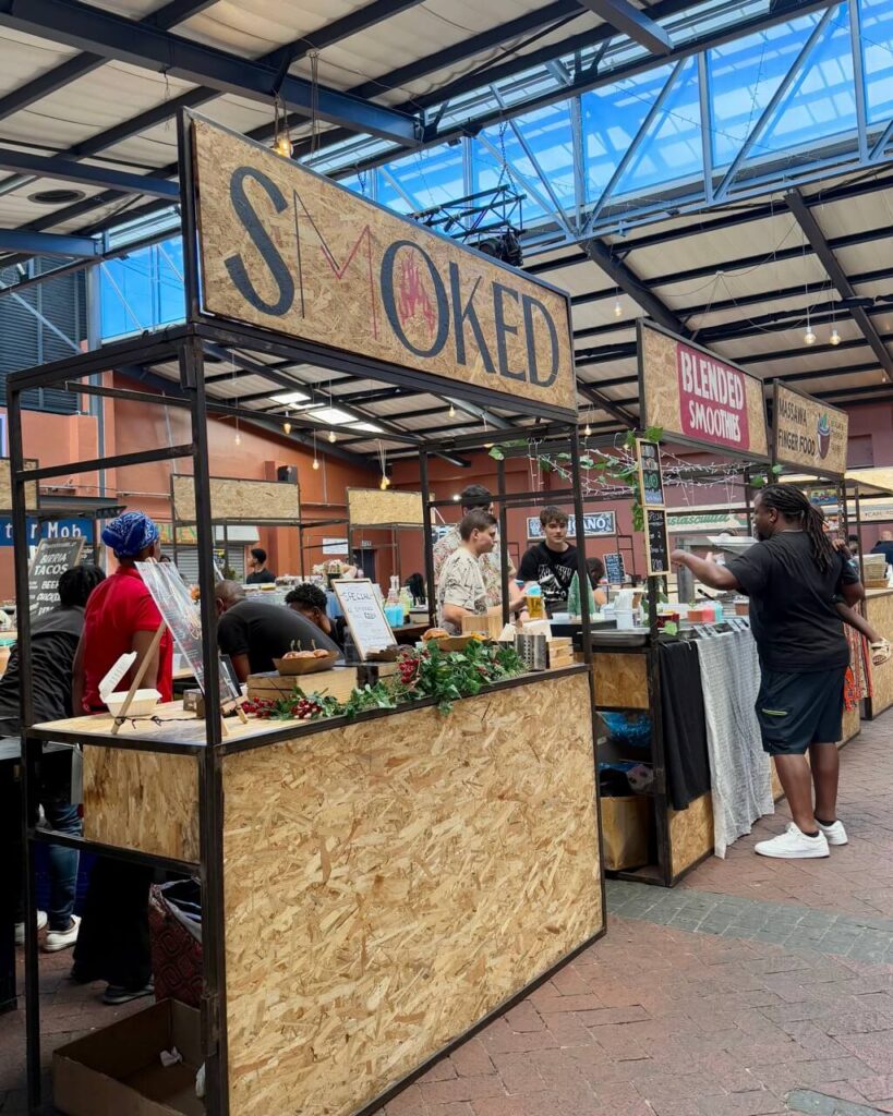 A lively urban courtyard at the Old Biscuit Mill featuring strings of hanging lights, brick buildings, and people dining at outdoor tables—blending industrial chic with Cape Town creativity