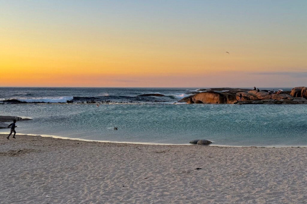 A rocky coastline cradles the Camps Bay tidal pool where people lounge and wade, surrounded by dramatic waves crashing nearby during the evening golden hour—nature and relaxation all in one.