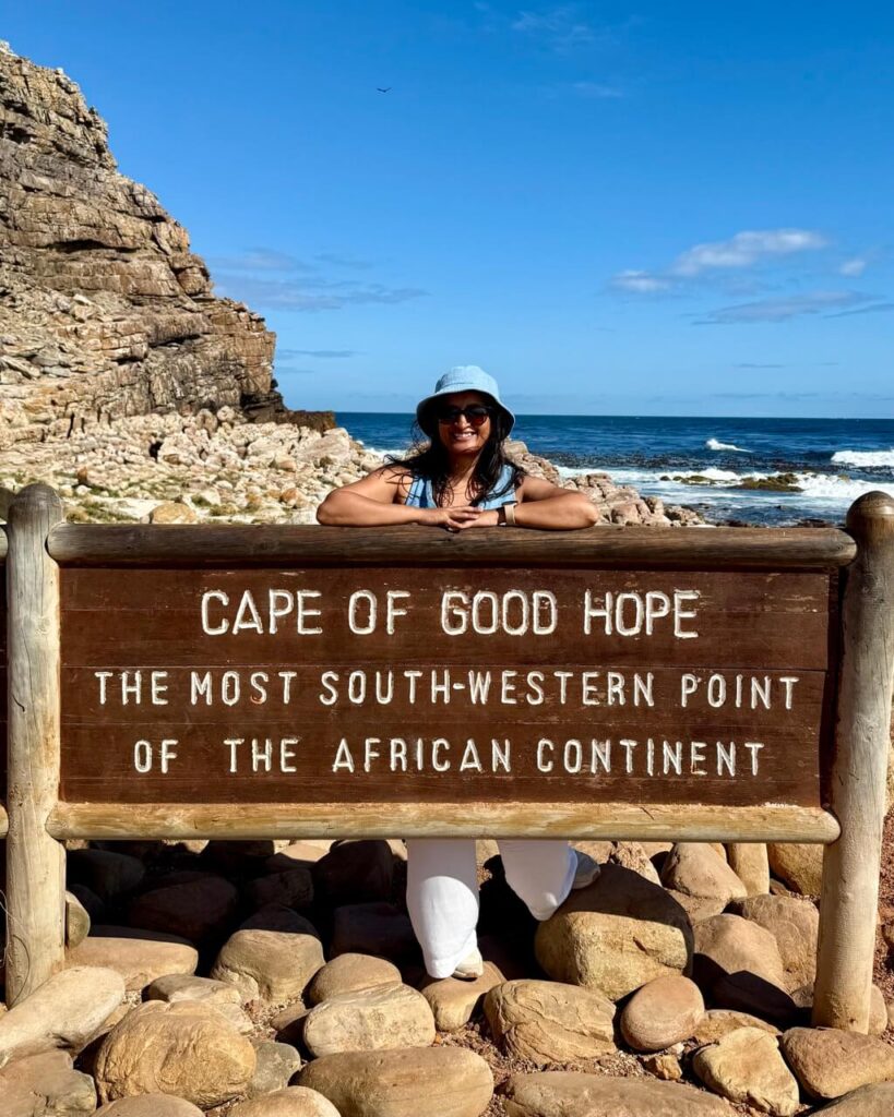 A scenic overlook at the Cape of Good Hope with dramatic cliffs, deep blue waters, and woman standing behind a wooden sign marking the iconic location, surrounded by wild, natural beauty.