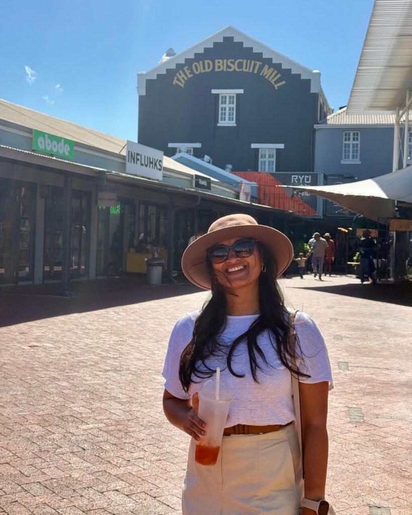 A woman in a white shirt and beige hat stands holding a drink in front of The Old Biscuit Mill market on a sunny day.
