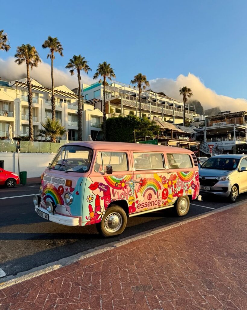 A colorful van decorated with bold patterns and "Cape Town" text is parked along a street in Camps Bay with palm trees and blue sky in the background, giving off a laid-back coastal vibe.
