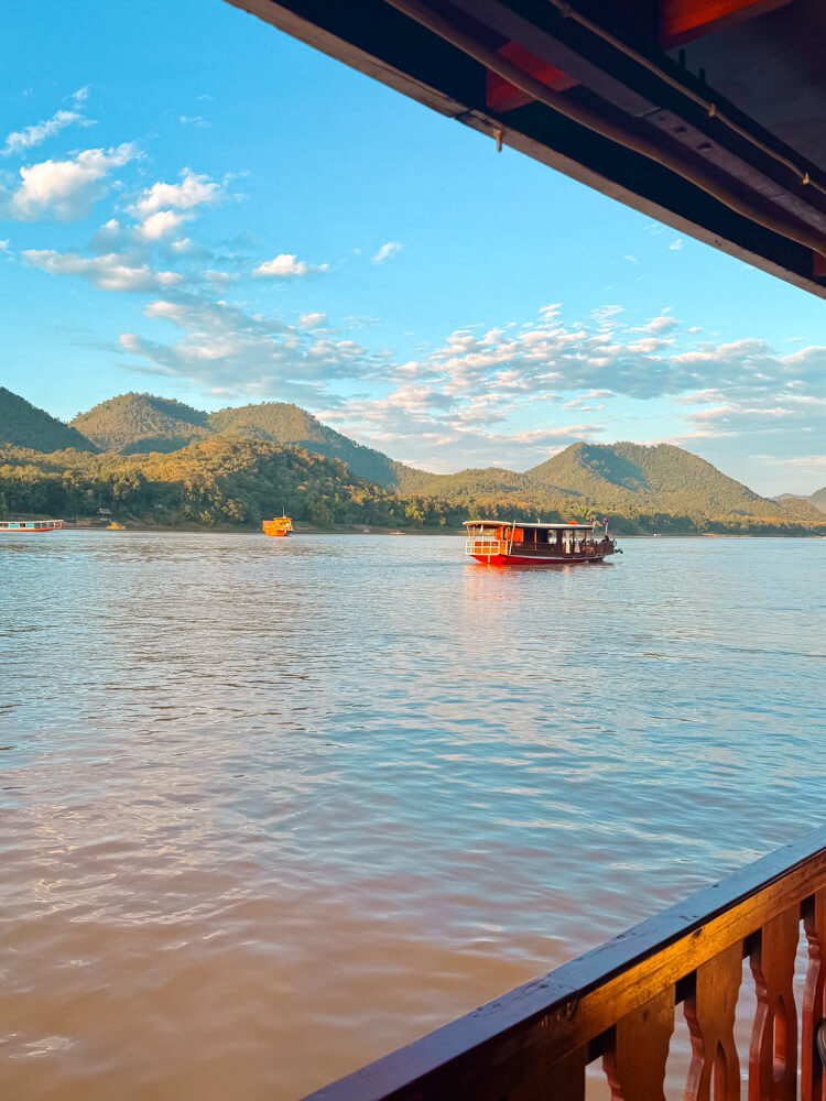 The Only Laos Itinerary You Need for 1 Week in Laos 25 View of the rolling hills and a boat over blue water from the Mekong River Cruise at sunset