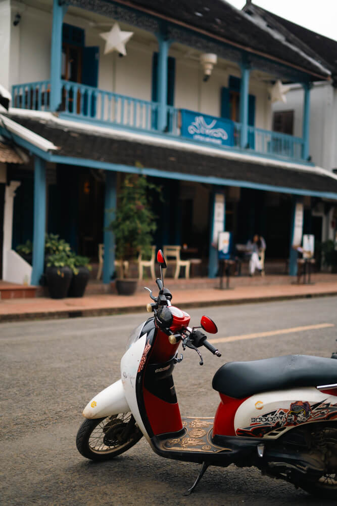 The Only Laos Itinerary You Need for 1 Week in Laos 4 A photo of a scooter on the road in Luang Prabang with the background of a blue and white colonial building