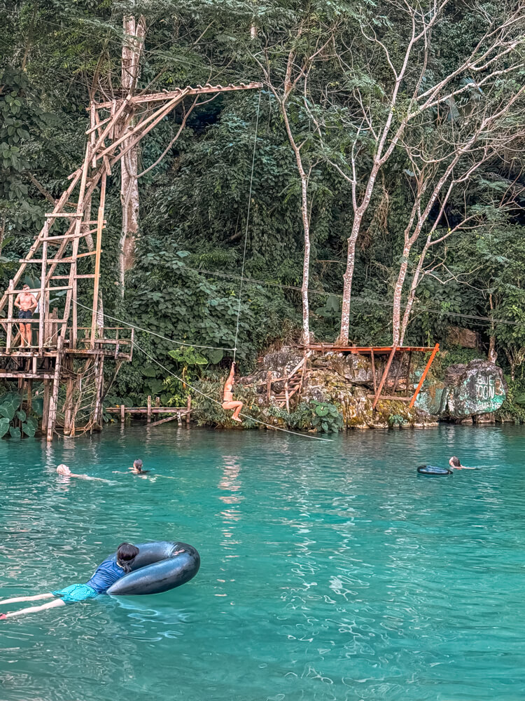 The Only Laos Itinerary You Need for 1 Week in Laos 14 A women hanging by a rope over the turquoise water of a lagoon ready to jump in while others are enjoying swimming and tubing into lagoon