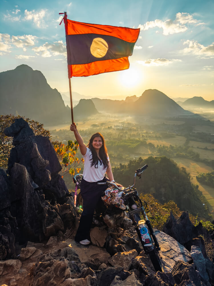 The Only Laos Itinerary You Need for 1 Week in Laos 3 A girl holding a flag of Laos while sitting on a bike on a mountain top as the sun rises behind her