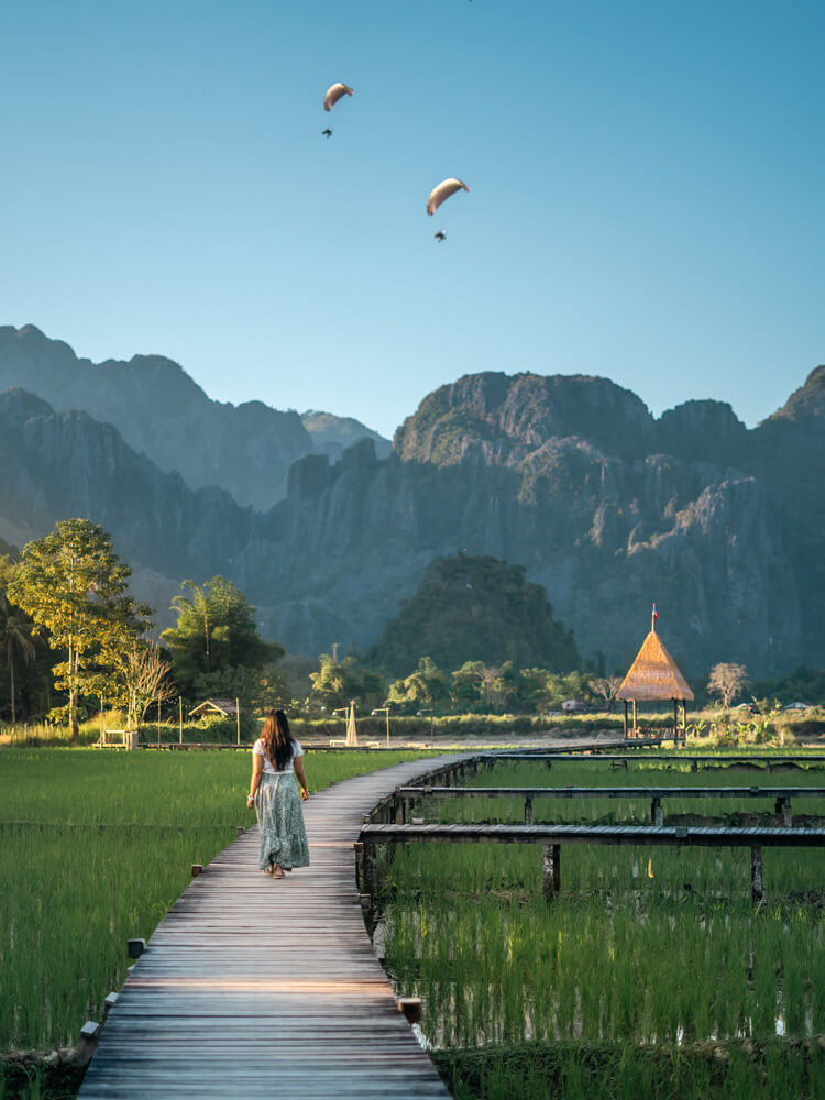 The Only Laos Itinerary You Need for 1 Week in Laos 11 A girl walking on a wooden walkway built over paddy fields with a view of jagged mountains and 2 paramotors in the air