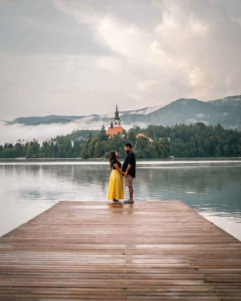 2-Day Lake Bled Itinerary: The Perfect Road Trip Guide 4 A couple looking at each other while holding hands at the dreamy lake Bled that has the church, mountains, and mist in the background