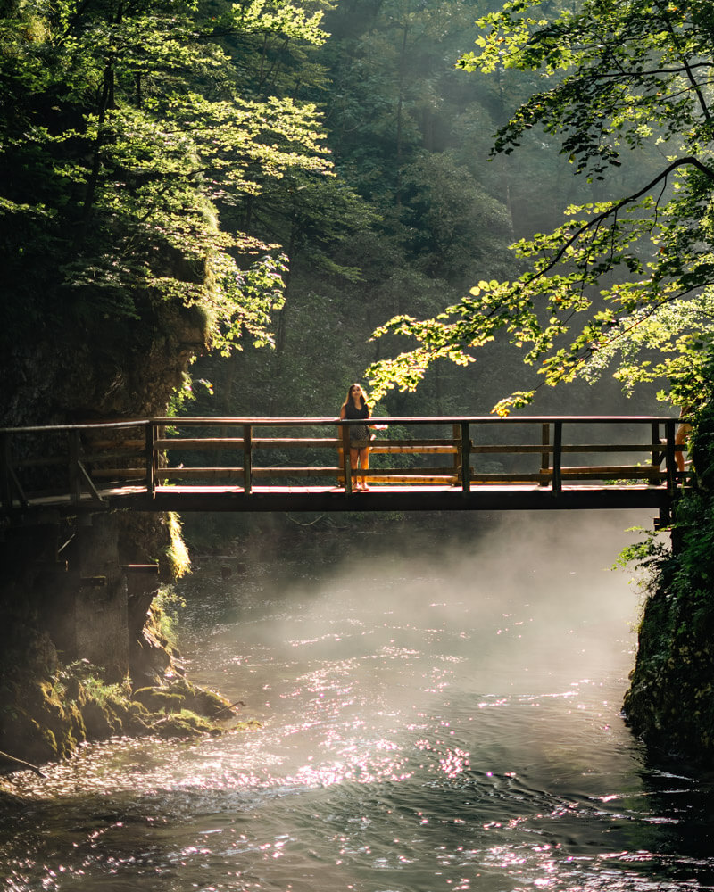 2-Day Lake Bled Itinerary: The Perfect Road Trip Guide 15 A girl standing on a narrow wooden bridge at Vingtar Gorge. It is an etheral scene with water below and mist with sunlight shining through