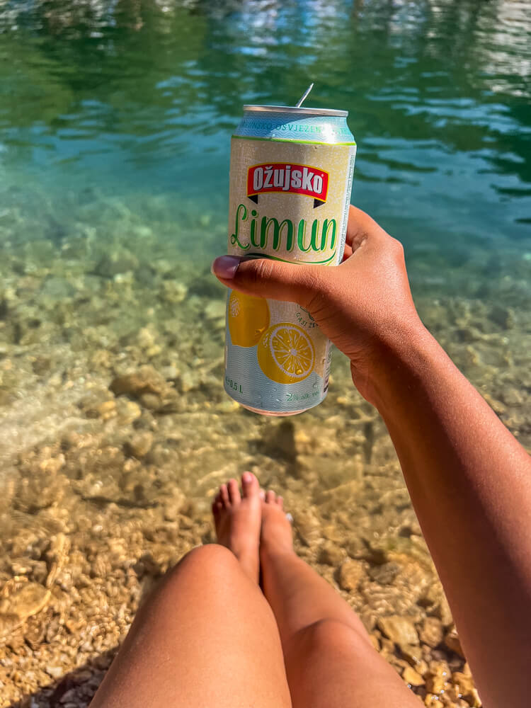 Day Trip to Mostar from Split or Dubrovnik (How to Plan It) 20 A girl sitting by the water posing with a canned drink -Ozujsko Limun