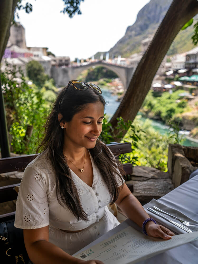Day Trip to Mostar from Split or Dubrovnik (How to Plan It) 16 A girl is sitting in a restaurant looking at the menu with a view of Stari Most bridge in the background