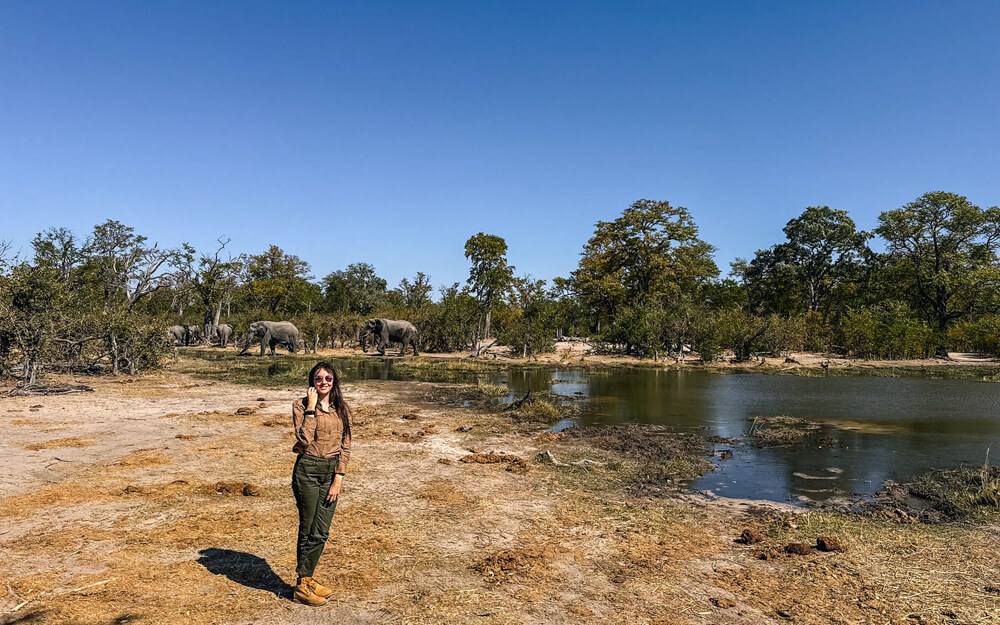 A girl poses by a water body with elephants in Okavango, Botswana.