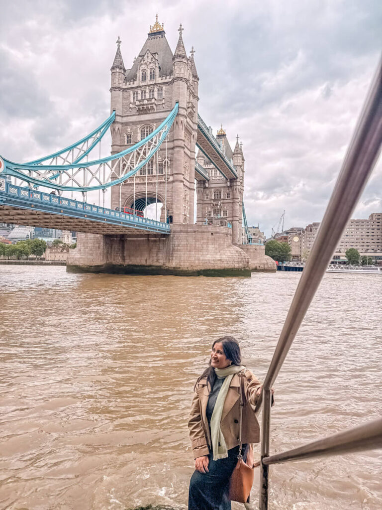 4-Day London Itinerary: Best of London + Easy Walking Routes 11 A woman in a beige coat smiles on a boat on the River Thames, with Tower Bridge's stone towers and blue suspension cables rising behind her.