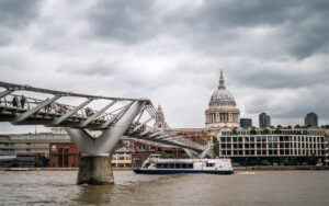 A view of the Millennium Bridge over the Thames as a boat goes by underneath and St. Pauls Church in the background on a cloudy moody day
