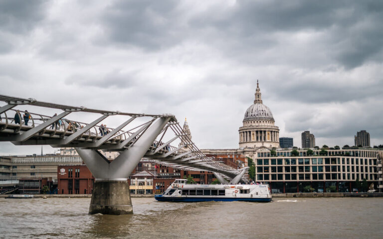 A view of the Millennium Bridge over the Thames as a boat goes by underneath and St. Pauls Church in the background on a cloudy moody day