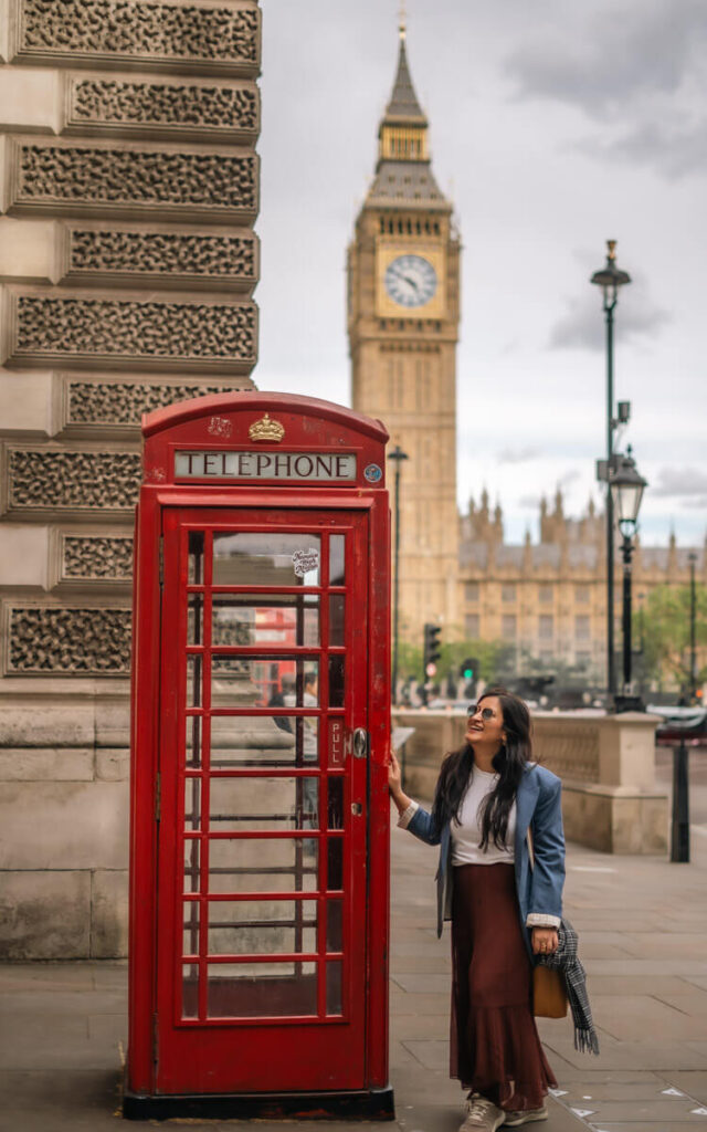 4-Day London Itinerary: Best of London + Easy Walking Routes 3 A girl wearing a brown skirt and blue blazer poses with a red telephone booth in London with the Elizabeth Tower in the backgroup