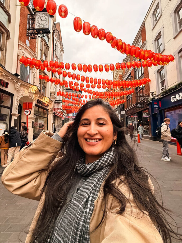 A smiling woman poses on a busy Chinatown London street strung with rows of red paper lanterns overhead.