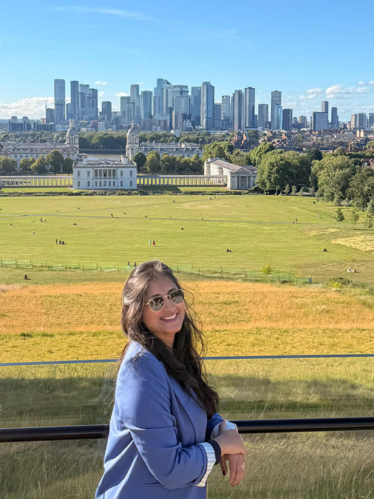 4-Day London Itinerary: Best of London + Easy Walking Routes 19 A woman in a blue blazer and sunglasses stands at a free viewpoint in Greenwich Park, with the Old Royal Naval College and Canary Wharf skyline visible across the green park below.