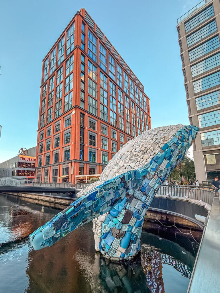 4-Day London Itinerary: Best of London + Easy Walking Routes 26 A large sculpture of a whale made from recycled blue and white plastic waste, rising out of the water at Canary Wharf with modern high-rise buildings behind it.