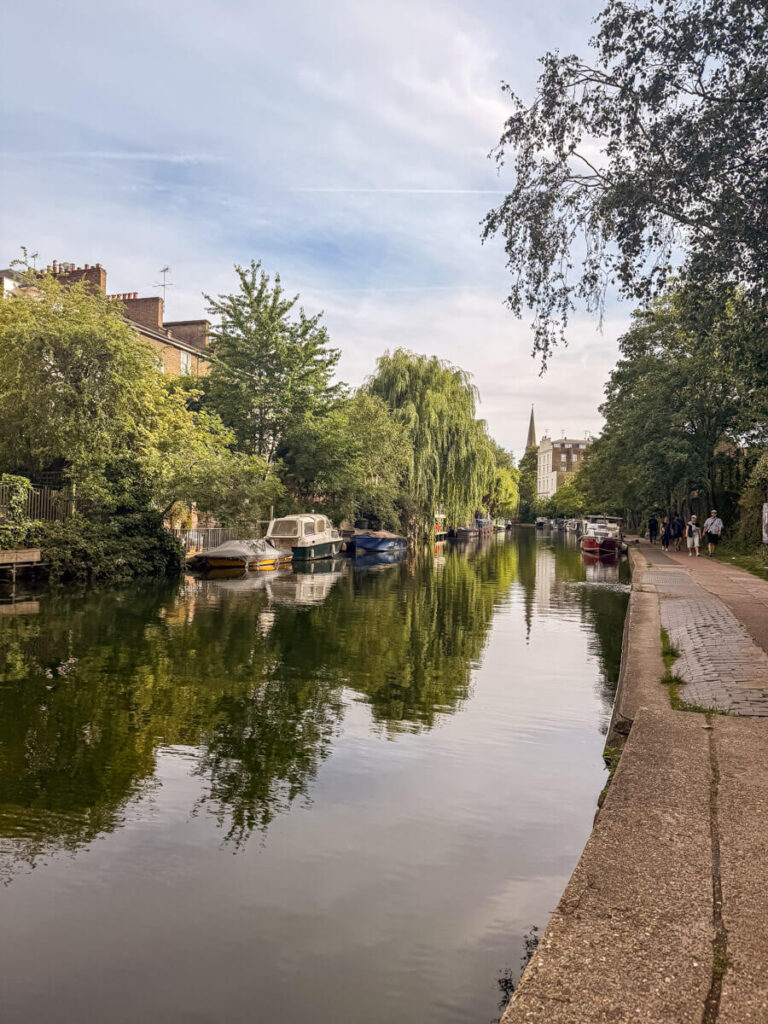 The Regent Canal in London is lined with weeping willows and houseboats, still water reflecting the trees, with a church spire peeking up in the distance and people walking the towpath.