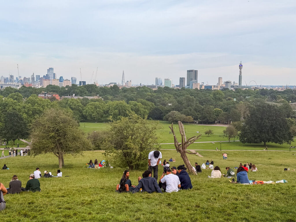 Crowds are relaxing on the grass at Primrose Hill with the city skyline, including the BT Tower, visible in the distance beyond the treeline.