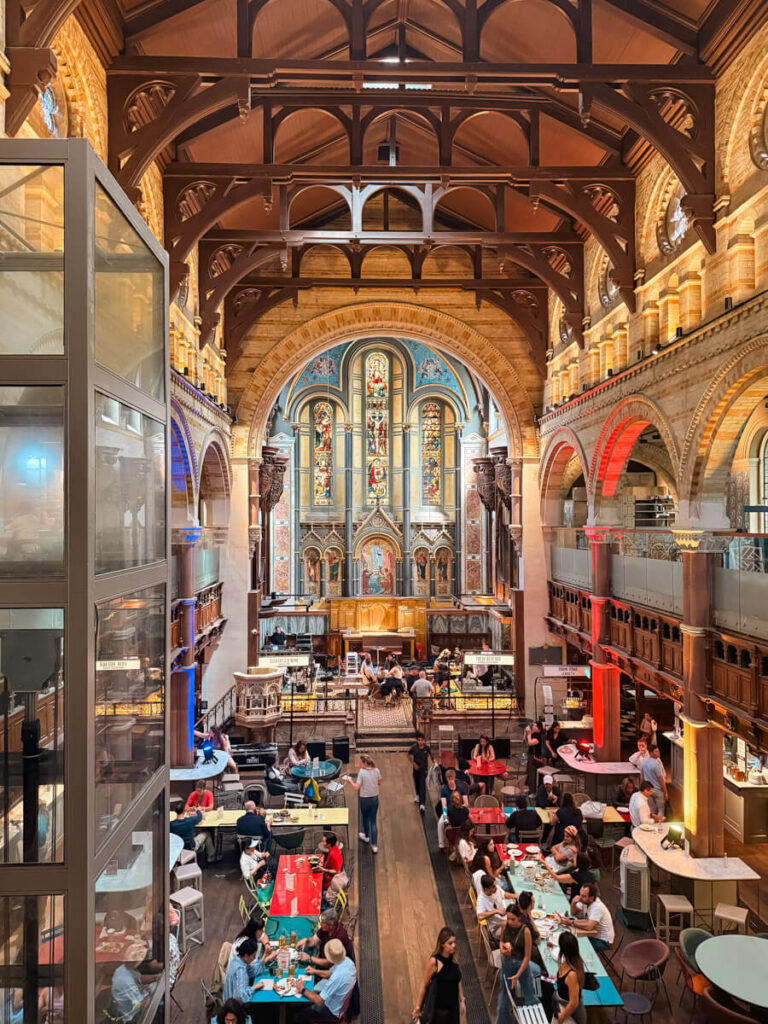 Marcato Mayfair is a former church turned into a lively food hall in London, with ornate stained glass and stone arches overhead and people eating at tables scattered across the wooden floor below.
